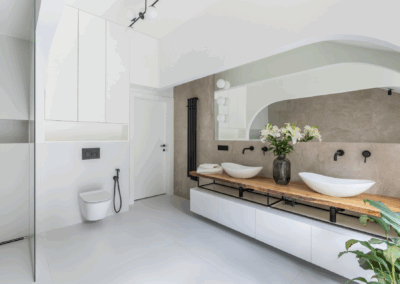 Strefa umywalek w minimalistycznej łazience z drewnianym blatem, białymi nablatowymi miskami i lustrem o zaokrąglonym kształcie.