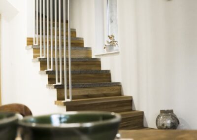 W tej realizacji każdy element ma swoje miejsce — także schody. Ich lekka konstrukcja, drewniane stopnie i pionowa balustrada tworzą formę, która nie dominuje, lecz płynnie wpisuje się w rytm wnętrza. Małe okno, ceramiczne detale i naturalne światło dodają przestrzeni miękkości. To projekt