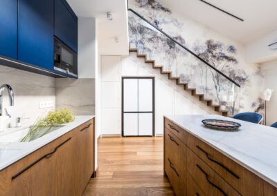 Aneks kuchenny z wyspą i schodami w apartamencie na Ursynowie — naturalne materiały, loftowe akcenty i ergonomiczny układ.