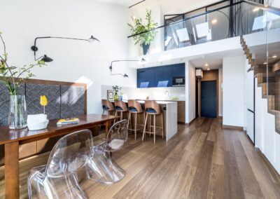 Strefa dzienna w apartamencie na Ursynowie — kuchnia z wyspą, jadalnia z ławką i schody na antresolę.